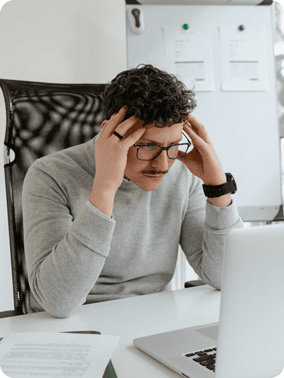A man with curly brown hair, glasses, and a mustache sits at a desk, looking down at a laptop with a stressed expression while holding his head in his hands.